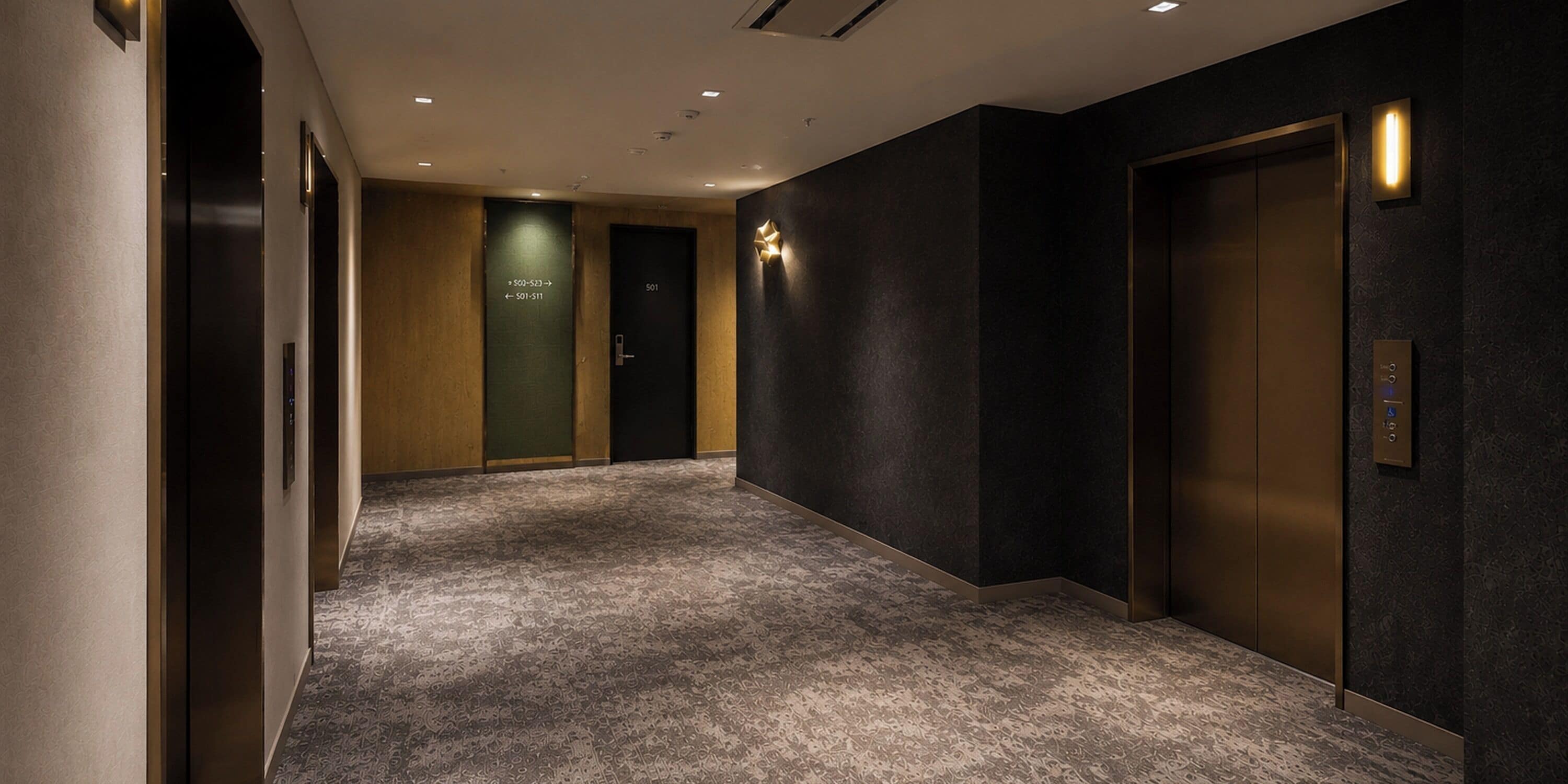 A hallway with a carpeted floor, a wall-mounted light fixture, and a door with a number.