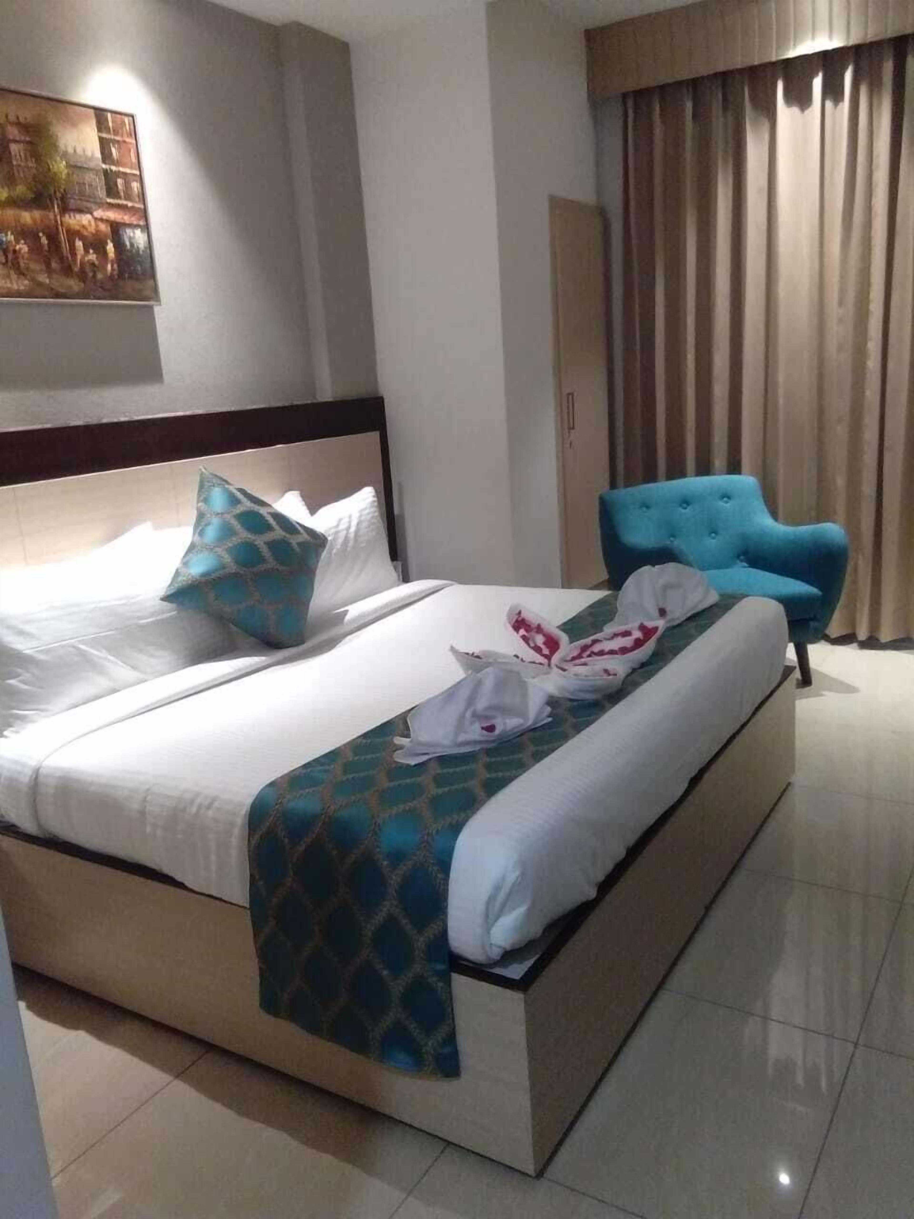 Quarto de hotel com uma cama grande, uma poltrona estofada azul e um quadro na parede.