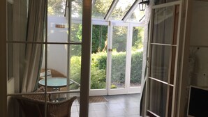 Una habitación con puerta de vidrio que da a un jardín, una silla de mimbre y una mesita.