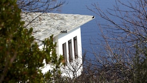 Un edificio de fachada blanca y techo inclinado, rodeado de árboles y con vista a un cuerpo de agua.
