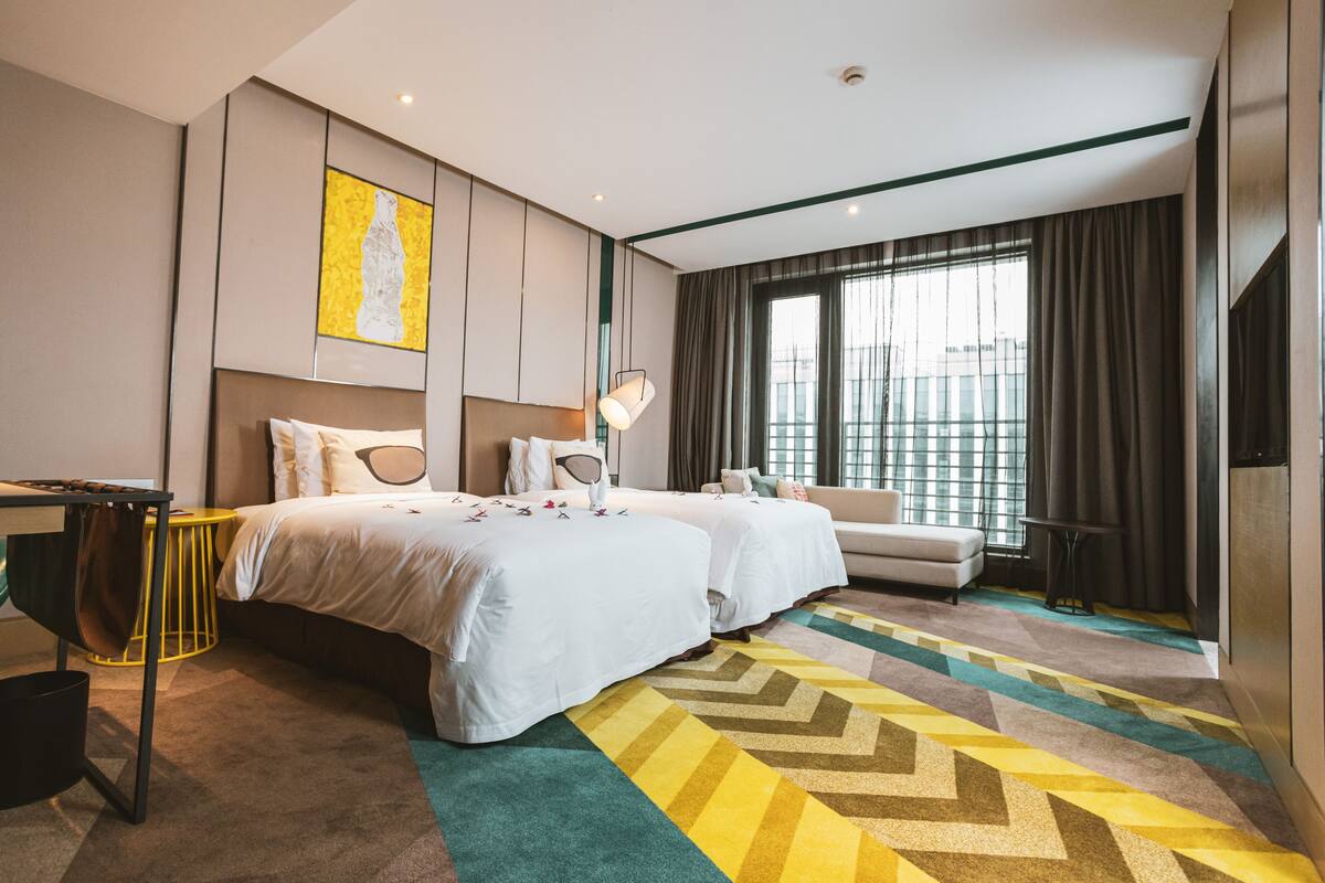 A hotel room with two beds, a sofa, a desk, and a large window with curtains.