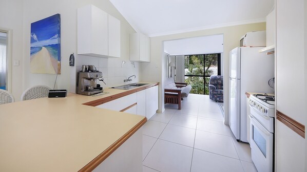 A modern kitchen with white cabinets, a coffee machine, and a stove. There is a dining area with a table and chairs visible through a glass door. - Quandamooka Beach House (Raceview)