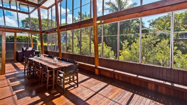 Un patio espacioso y cubierto con piso de madera, ventanas amplias y vista a exuberante vegetación.