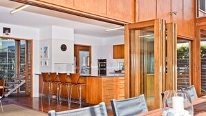 Una cocina moderna con armarios de madera, un comedor con mesa y sillas de madera, y vistas al exterior a través de amplios ventanales.