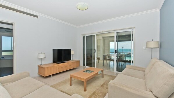 A modern living room with a flat-screen TV, wooden furniture, a glass door leading to a balcony with a view of the sea.