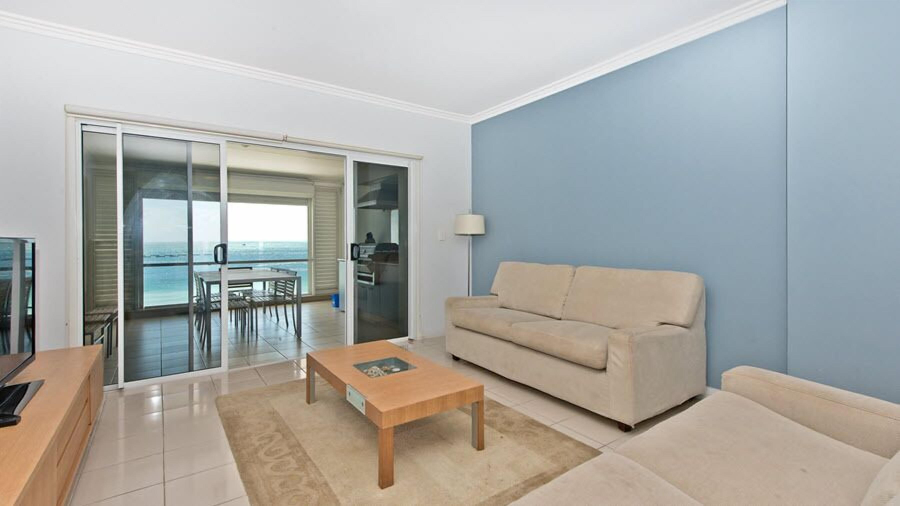 Una sala de estar moderna con un sofá, una mesa de centro y un televisor. Hay un balcón con vista al mar.