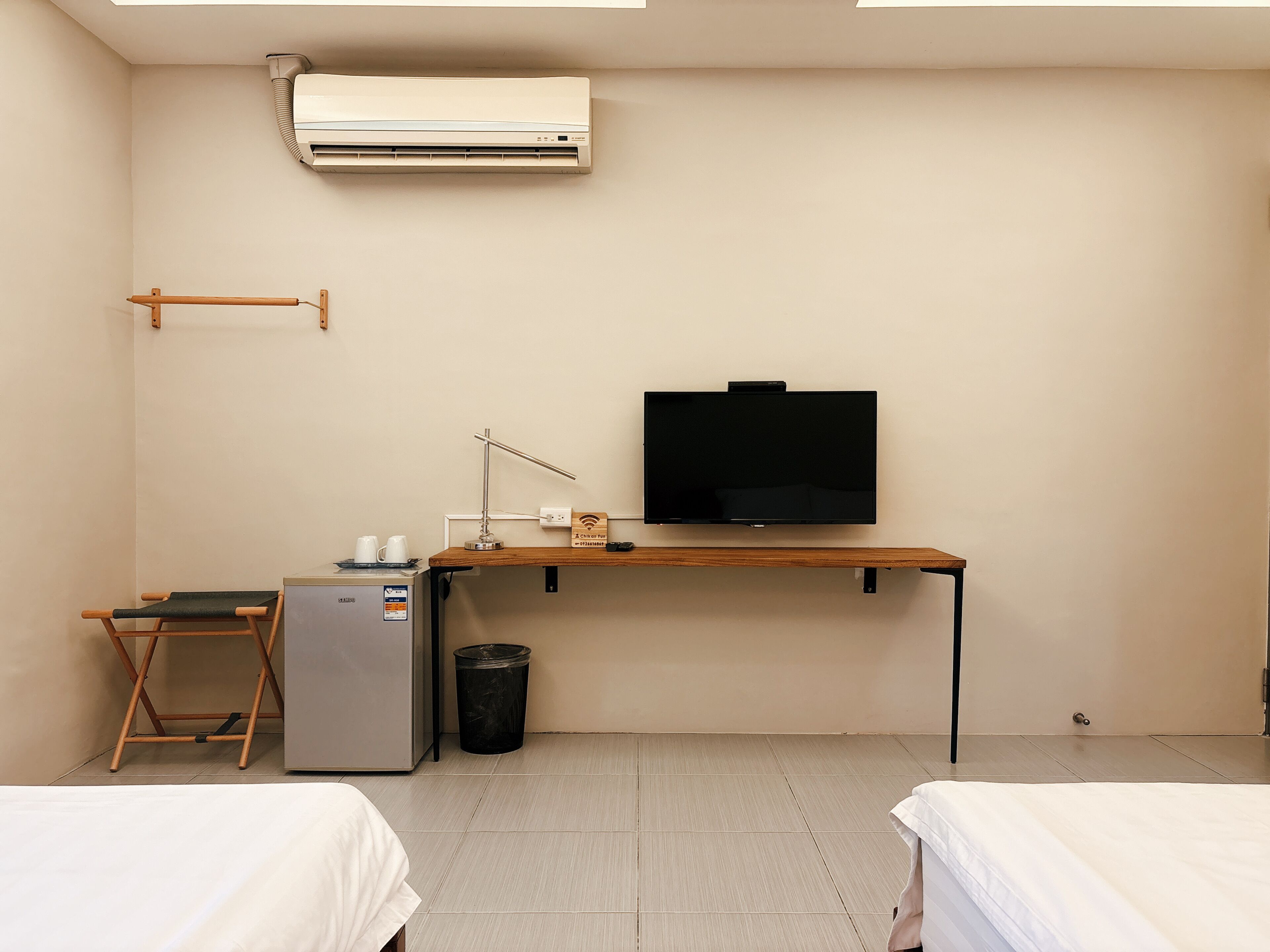 A hotel room with a bed, a desk with a TV, a small fridge, a trash bin, and an air conditioning unit.