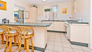 A kitchen with a central island, bar stools, a refrigerator, and a window with curtains. - Curlew Corner (Cowan Cowan)
