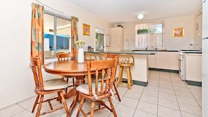 A dining area with a round wooden table and six chairs, a kitchen with white cabinets, and a window with curtains. - Curlew Corner (Cowan Cowan)