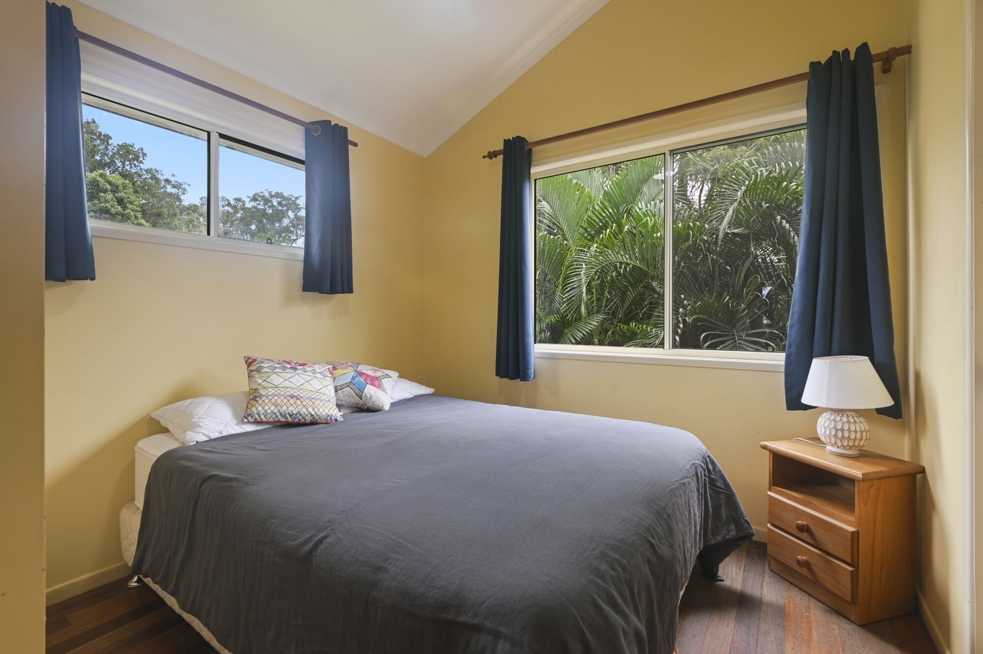 Un dormitorio con una cama grande, dos almohadas, una mesita de noche con una lámpara y una ventana con cortinas.