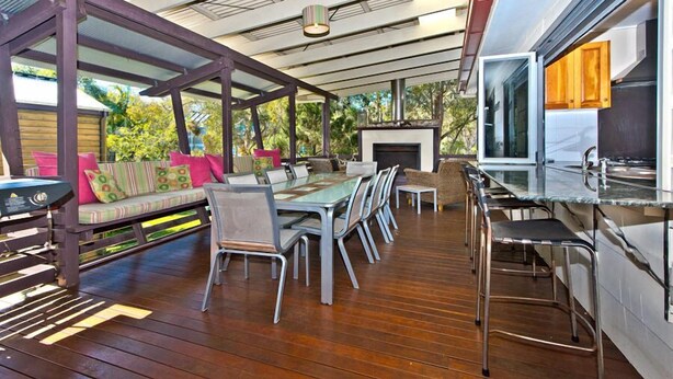 A spacious outdoor dining area with a long table and chairs, a built-in grill, and a cozy seating area with cushions. - Moreton Magic (Cowan Cowan)