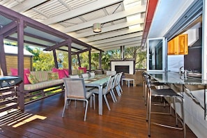 Un vaste espace extérieur aménagé pour les repas, avec une longue table et des chaises, un barbecue intégré et un coin salon confortable agrémenté de coussins.