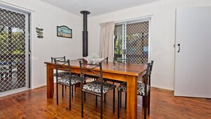 A dining area with a wooden table and chairs, a black chimney, and a view through a window with a curtain. - Moreton Magic (Cowan Cowan)