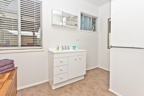 Un baño con tocador blanco, un espejo, una ventana con persianas y un banco de madera.