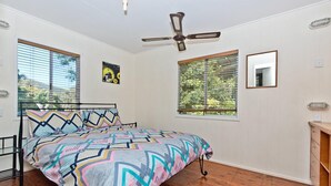 A bedroom with a bed, a ceiling fan, two windows with blinds, and a small mirror on the wall. - Moreton Magic (Cowan Cowan)