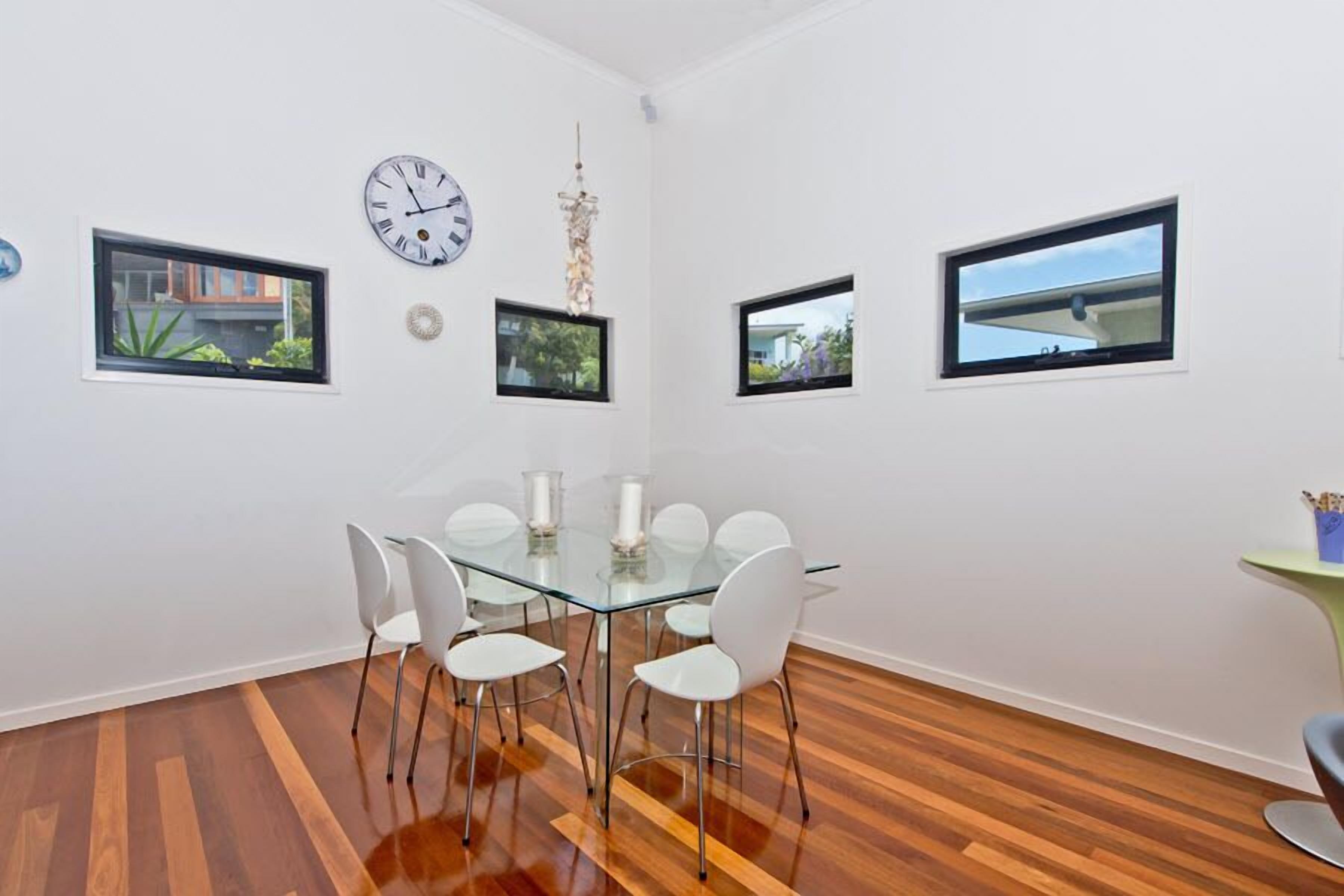 Comedor moderno con mesa de vidrio, sillas blancas y piso de madera. Se observan tres ventanas con vistas a edificios y zonas verdes.