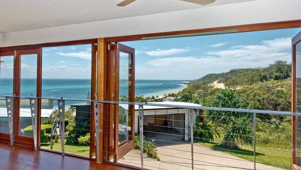 A balcony with a glass door leading to a scenic ocean view, a wooden floor, and a clear sky.