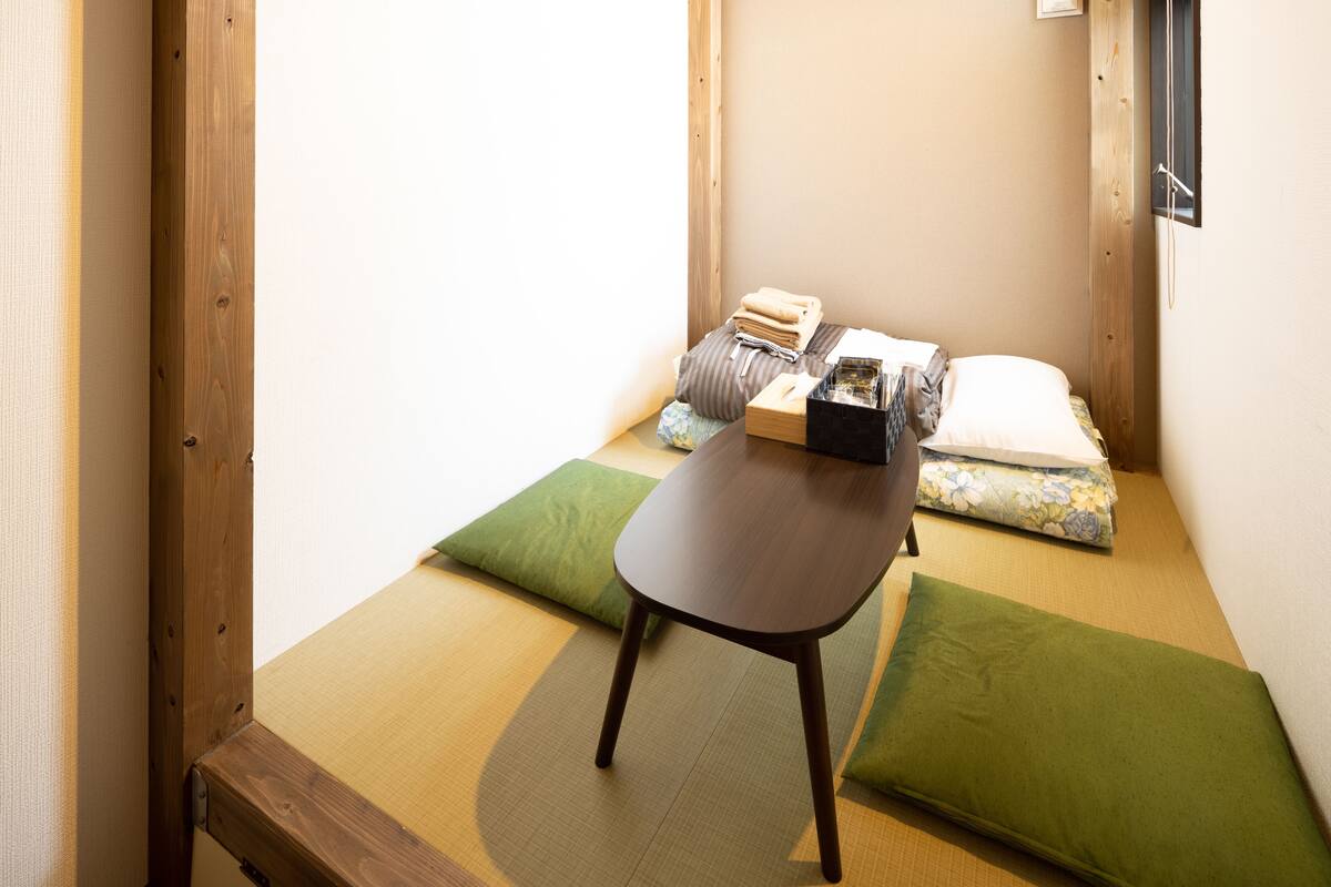 Ein Raum mit Tatami-Matten, einem niedrigen Holztisch, grünen Kissen und einem Bett mit einer floralen Decke.