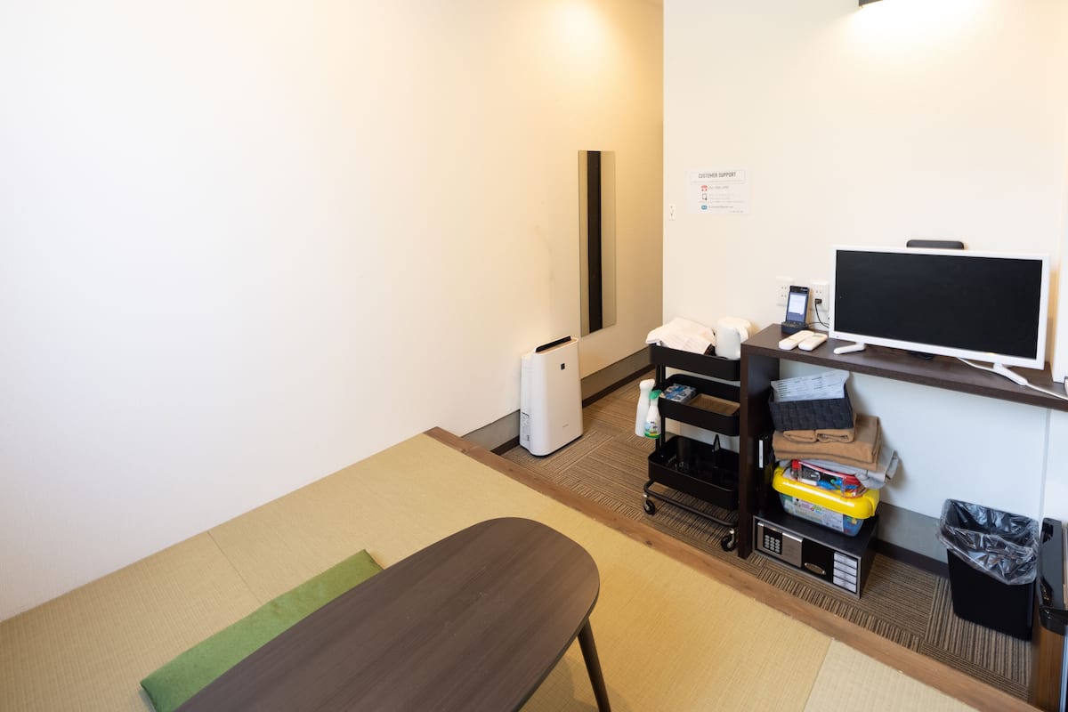 Ein Hotelzimmer mit einem Bett, einem Schreibtisch mit Computer, einem Fernseher, einem Heizkörper, einem Mülleimer und einem kleinen Regal mit Gegenständen.