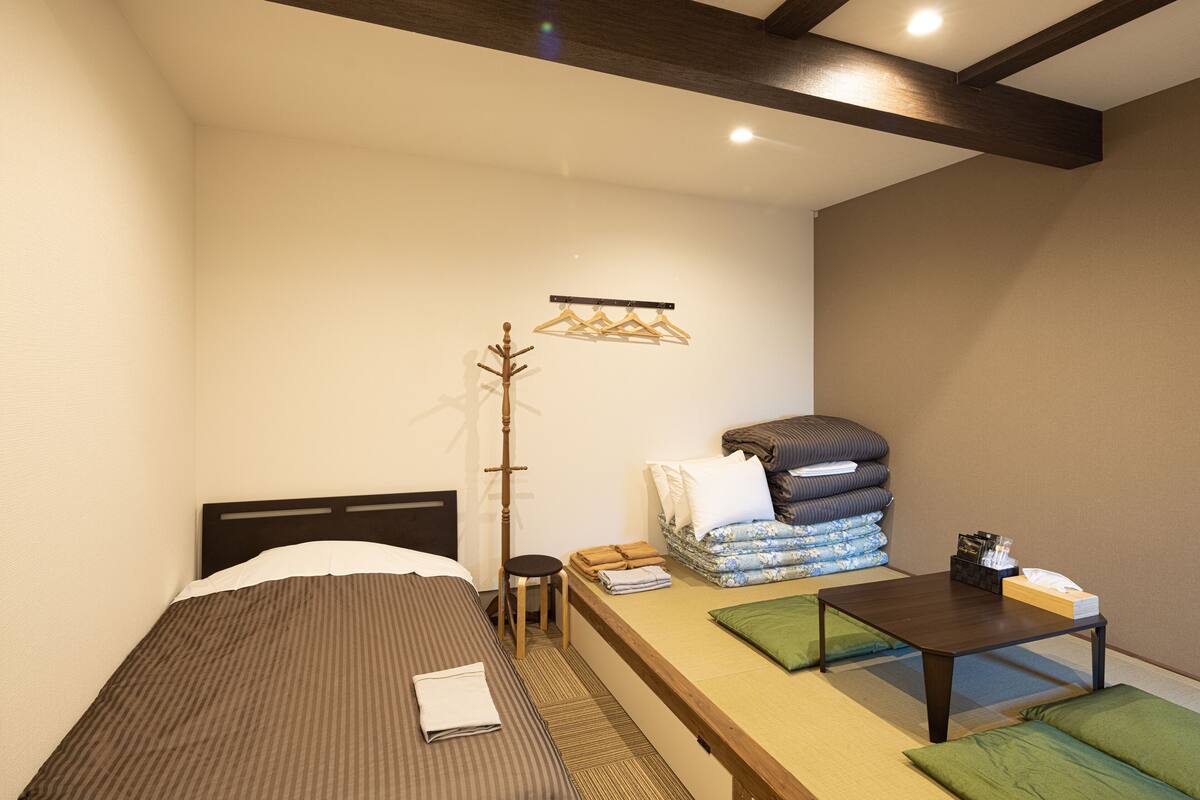 Ein kleines Zimmer mit einem Einzelbett, einem kleinen Tisch mit Taschentuchbox, einem Stuhl und einem Wandhaken für Kleidung.