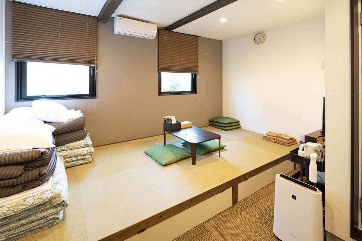 Ein Zimmer mit Tatami-Matten, einem Bett mit Kissen, einem Tisch mit Sitzkissen, einer Wanduhr und einem kleinen Kühlschrank.