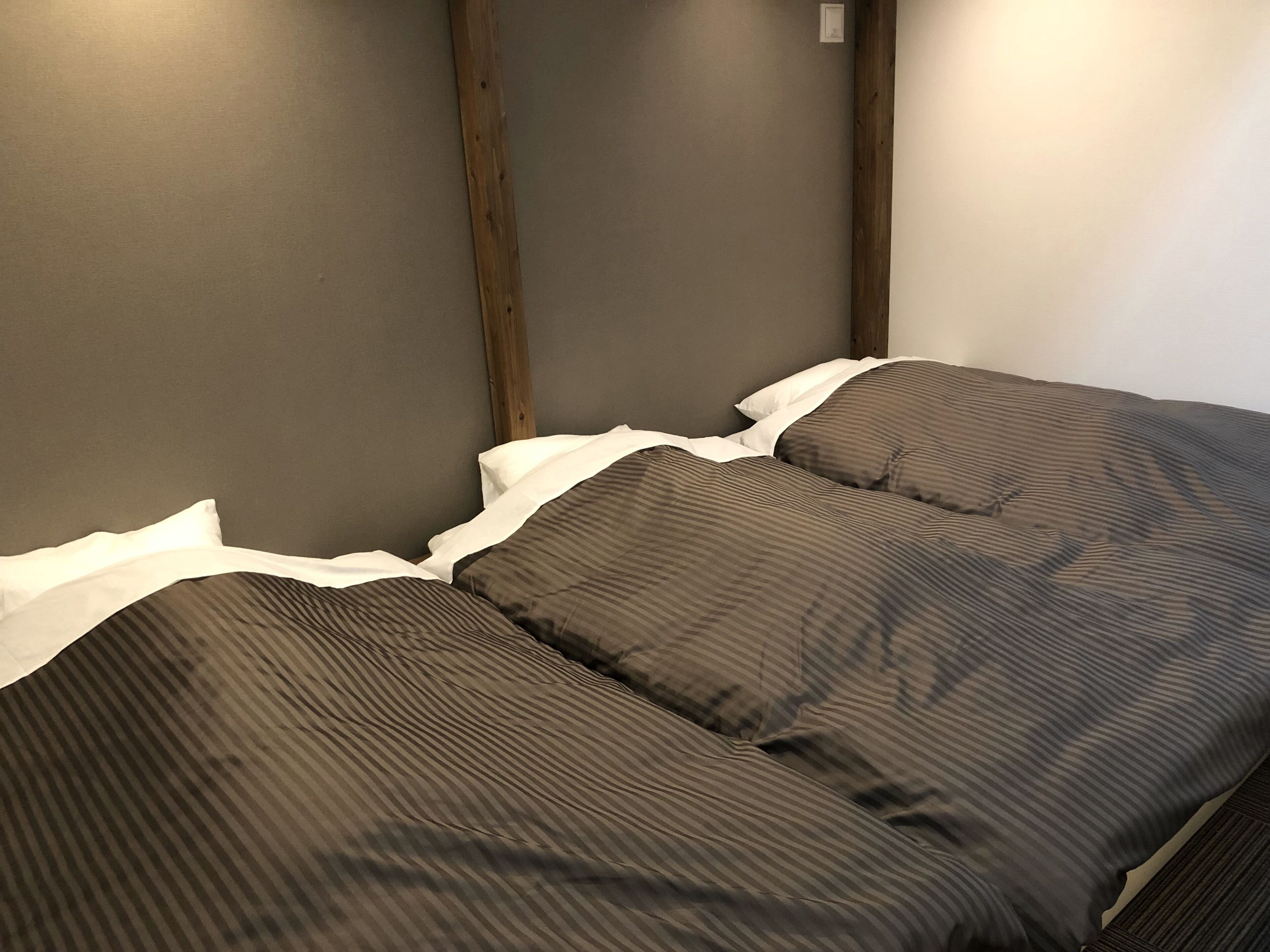 Una cama individual con una colcha a rayas y un almohadón blanco, frente a una pared lisa con una viga de madera.