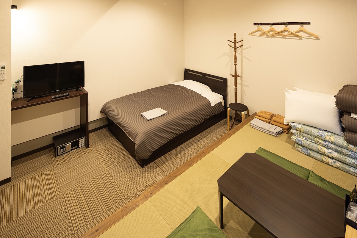 A hotel room with a bed, a TV on a stand, a bedside table, a chair, and a coat rack.