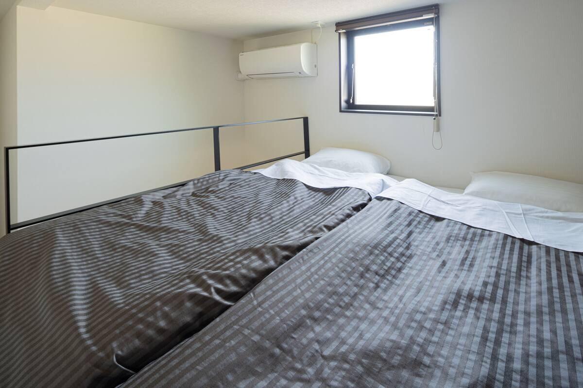 Ein Einzelbett mit Metallgestell, ein Fenster mit Jalousien und eine Klimaanlage an der Wand.