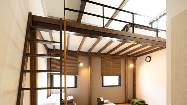 Un dormitorio estilo loft con techo de viga de madera, dos ventanas con persianas, una cama, una mesita y un cojín verde.