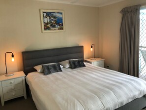 A neatly made bed with a striped duvet, two bedside tables with lamps, a painting on the wall, and a window with curtains. - Villa at Tangalooma - Villa 14 (Moreton Island)