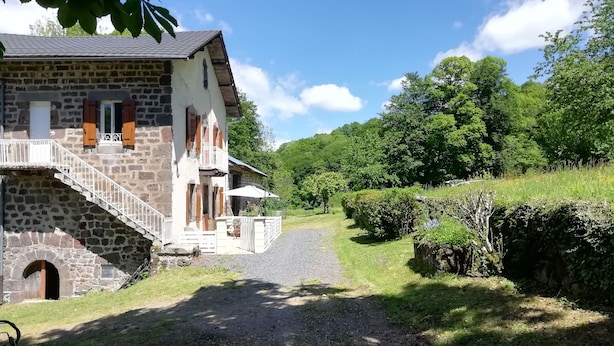 Ein Steinhaus mit einer Treppe, die zu einem Balkon führt, umgeben von üppiger Vegetation und einem Kiesweg.