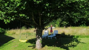 Ein Baum mit einer Schaukel, zwei weiße Stühle und ein Grill unter dem Baum.