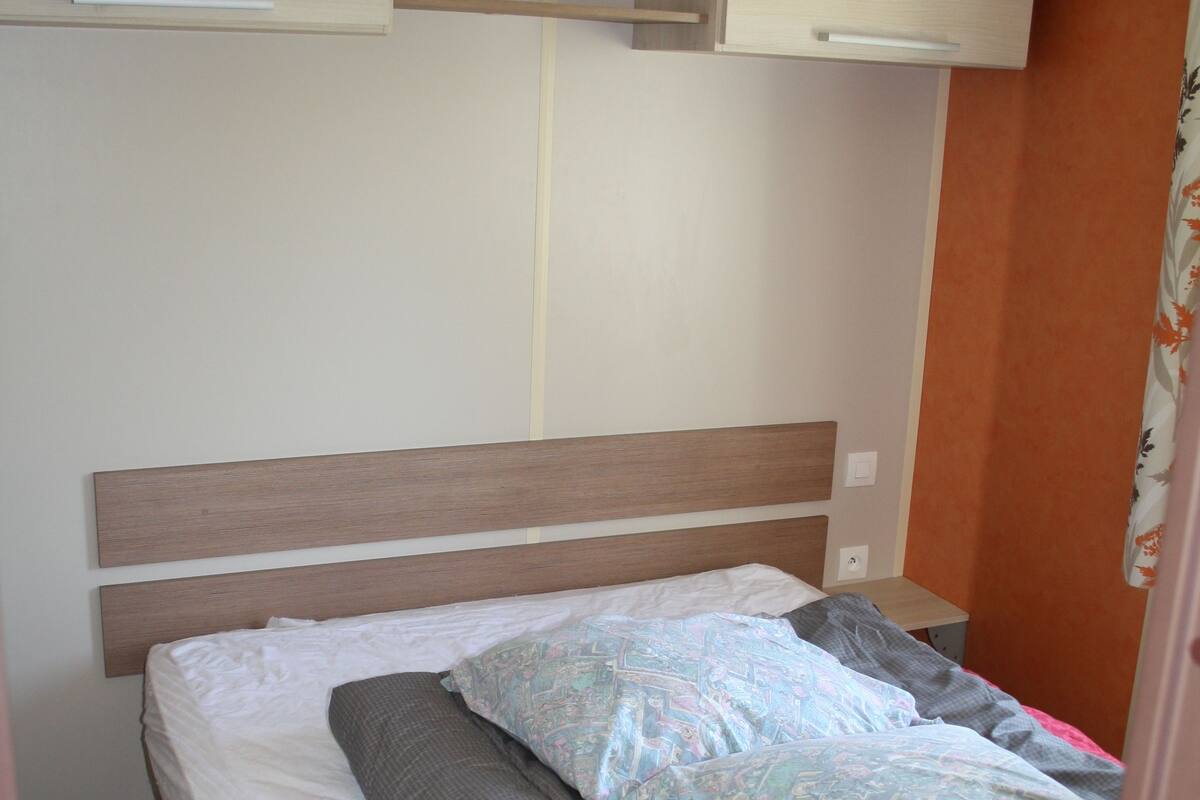 Een eenpersoonsbed met een houten hoofdbord, een klein nachtkastje en een inbouwplank boven het bed.