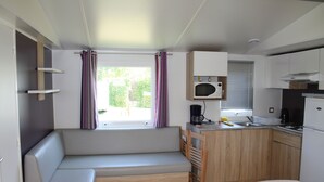 Een kleine keuken met een magnetron, fornuis en spoelbak. Er is een houten bank met opbergladen en een raam met gordijnen.