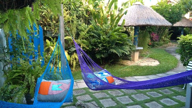 A hammock in a lush garden with a thatched-roof structure in the background. - House - Mar Condominium Azul (Porto de Galinhas)