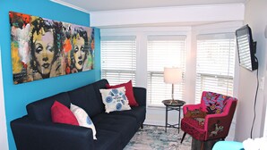 A living room with a dark blue sofa, red and blue pillows, a colorful painting, a small table with a lamp, and a red chair with a floral pattern. - Stylish Ocean Grove Apt - 4 Blocks from Beach & 2 Blocks from Downtown Asbury (Ocean Grove)