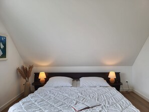 A bedroom with a large bed, two bedside lamps, a nightstand, a vase with dried plants, and a painting on the wall.