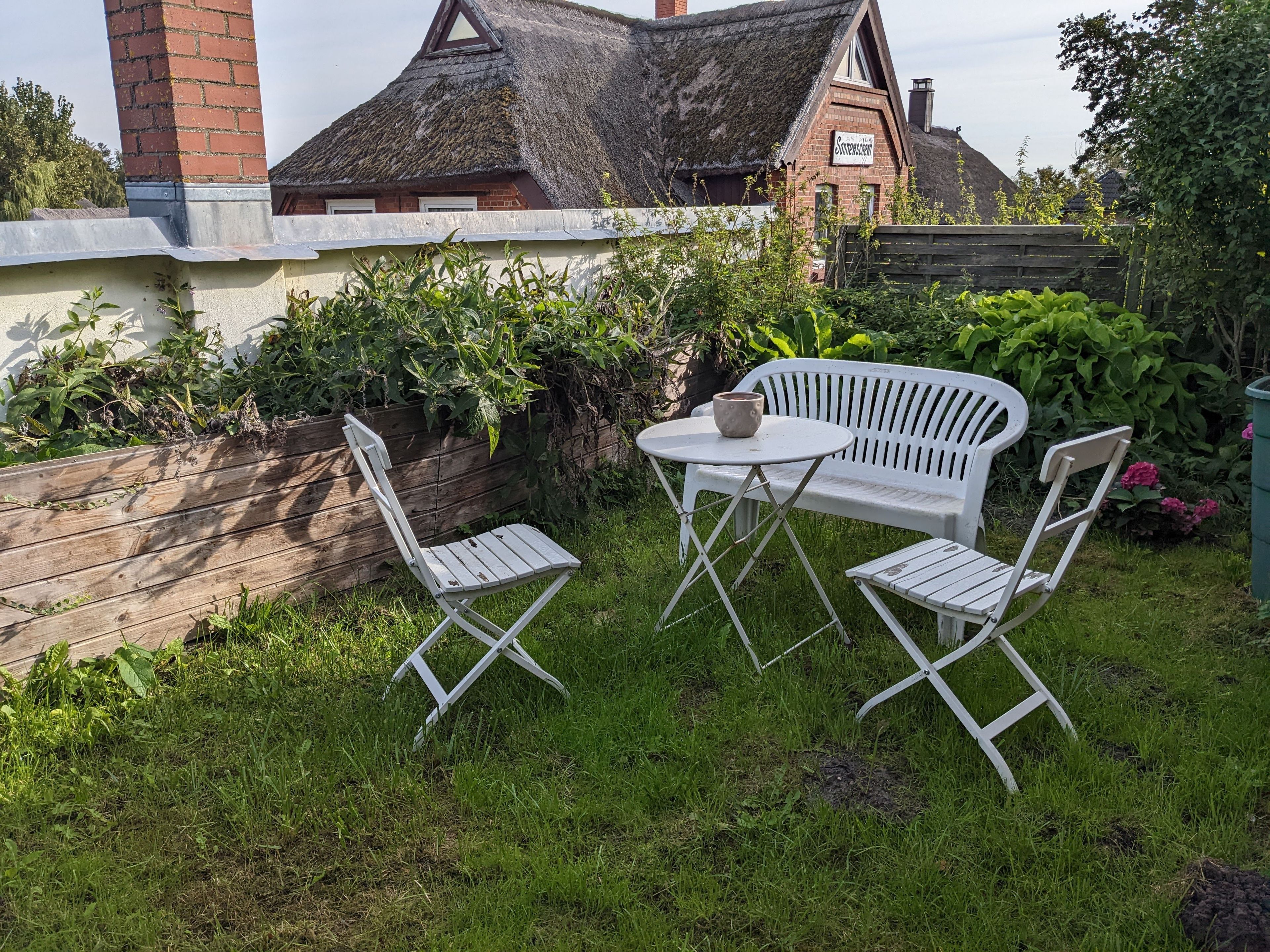A white metal bench and table set in a grassy garden area with a brick wall and a thatched-roof building in the background.