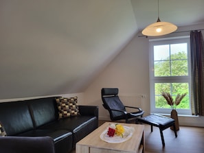 A living room with a black leather sofa, a black leather chair, a wooden coffee table with a vase of flowers, a hanging light fixture, and a large window with a view of greenery.