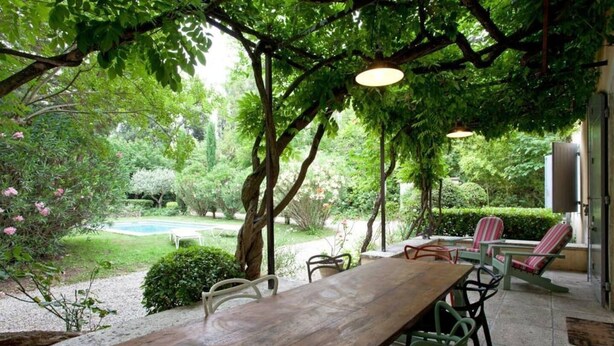 An outdoor patio with a wooden table and chairs, surrounded by lush greenery and trees.
