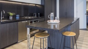 A modern kitchen with dark cabinetry, a black countertop, stainless steel appliances, and two wooden stools. - Avani Melbourne Box Hill Residences (Box Hill)