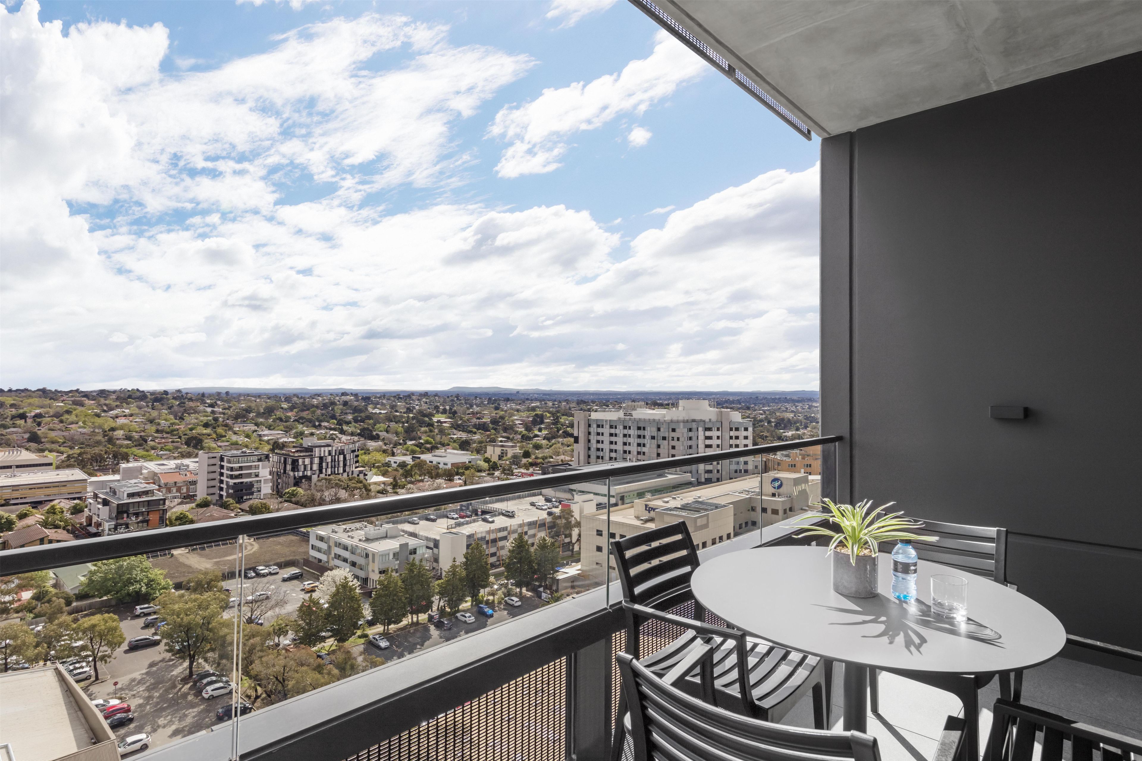 A balcony with a table and chairs, overlooking a cityscape with buildings and a clear sky.