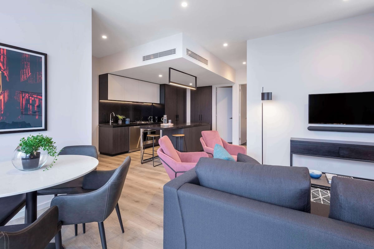 A modern living room with a grey sofa, a dining table with chairs, a kitchen area, and a flat-screen TV mounted on the wall.