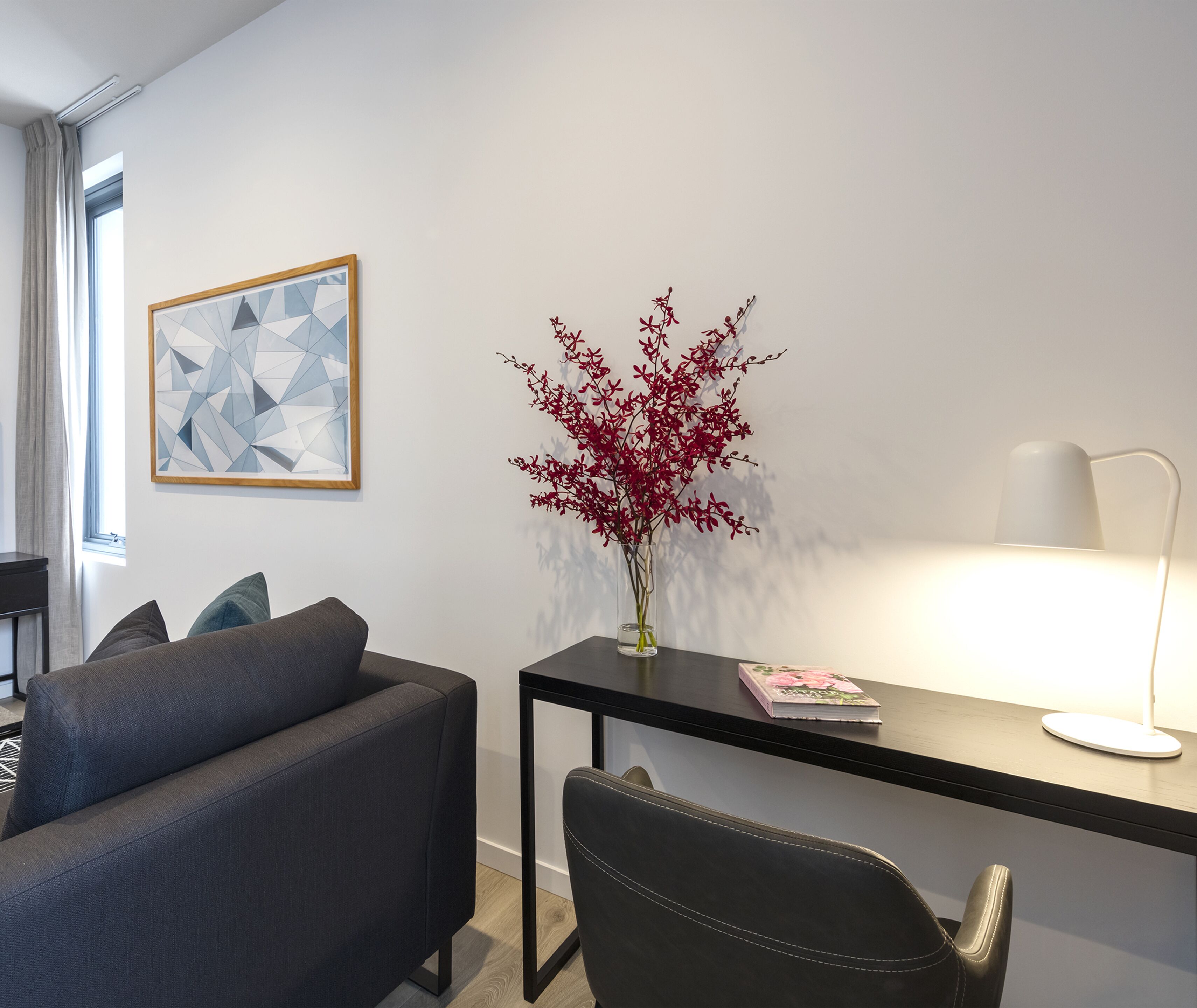 A modern room with a grey sofa, a black desk, a vase with red flowers, a framed geometric artwork, and a white lamp.