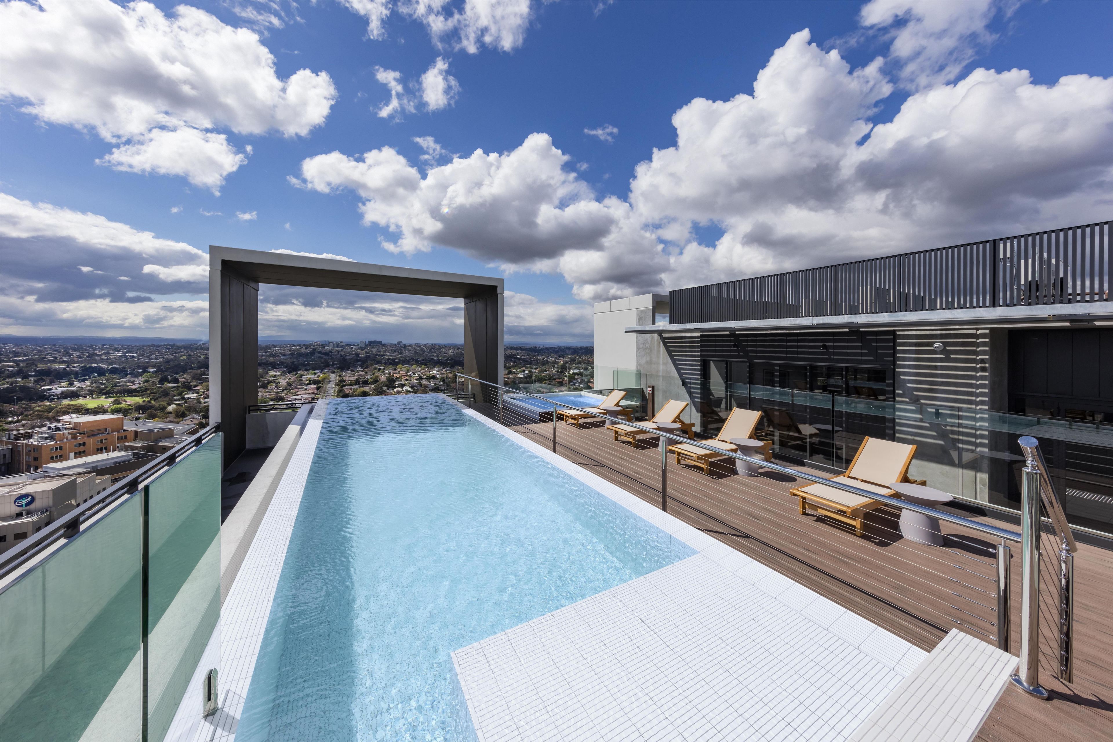 A rooftop infinity pool with a clear view of the cityscape, lounge chairs, and a modern building structure.