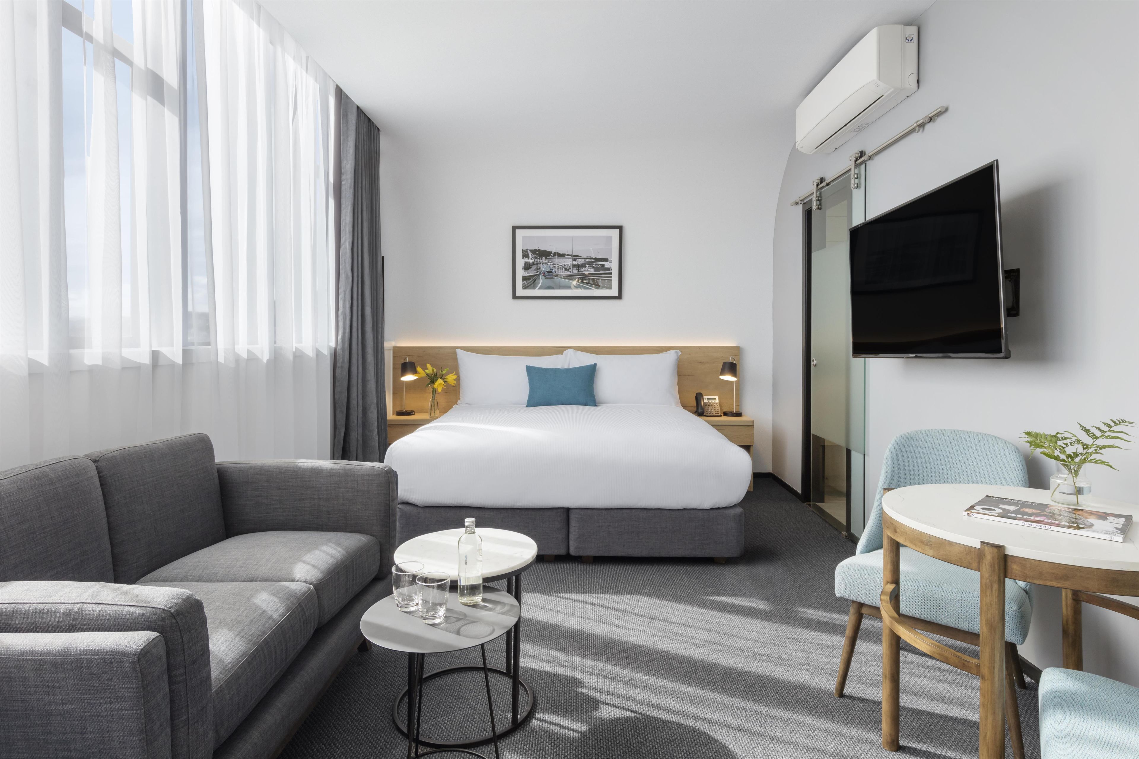 Quarto de hotel com cama, sofá, mesinha com copos, televisão e quadro na parede.