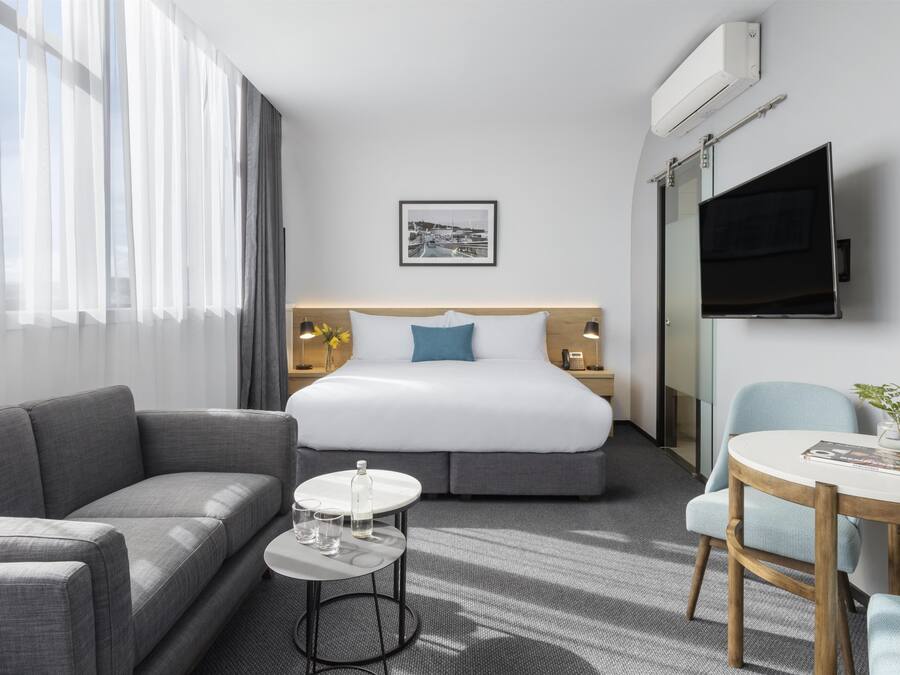 Habitación de hotel con cama, sofá, una mesita con vasos, televisor y un cuadro en la pared.