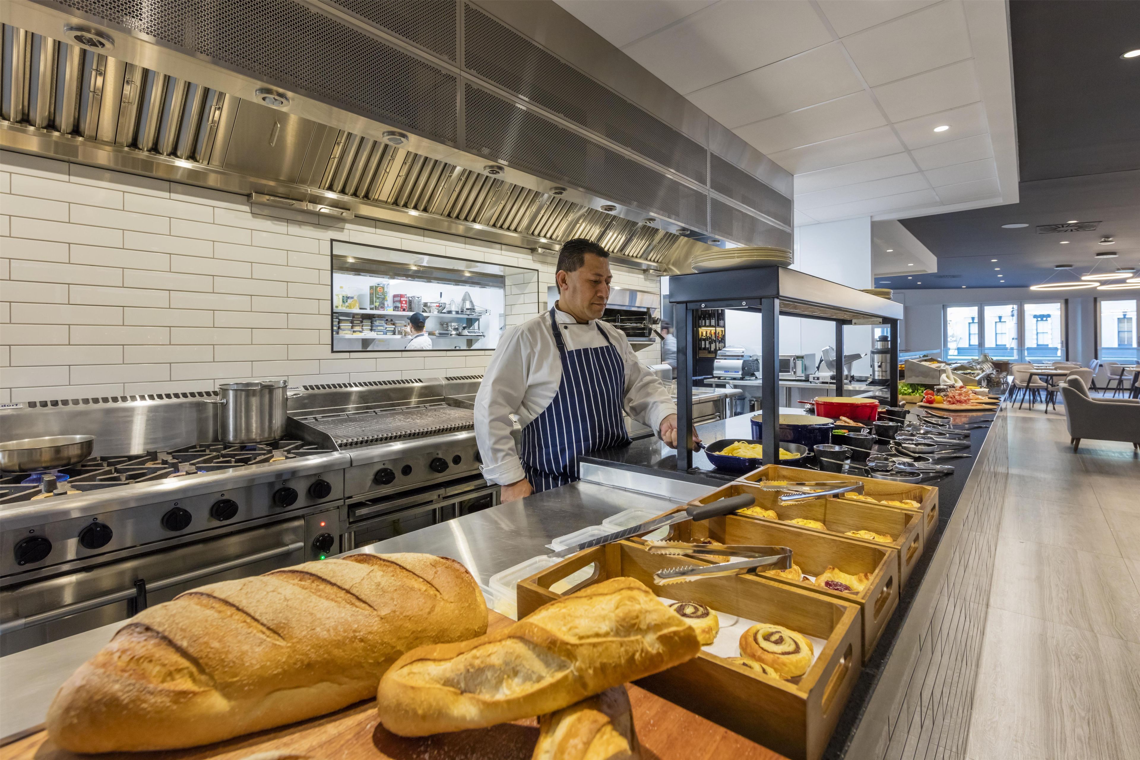 Cozinha comercial com eletrodomésticos de aço inoxidável, um chef de uniforme e uma mesa de buffet com pães e doces.