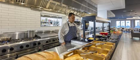 Una cucina professionale con elettrodomestici in acciaio inossidabile, uno chef in divisa e un'area buffet con pane e dolci.