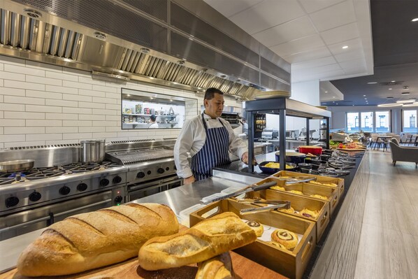A commercial kitchen with stainless steel appliances, a chef in uniform, and a buffet line with bread and pastries.